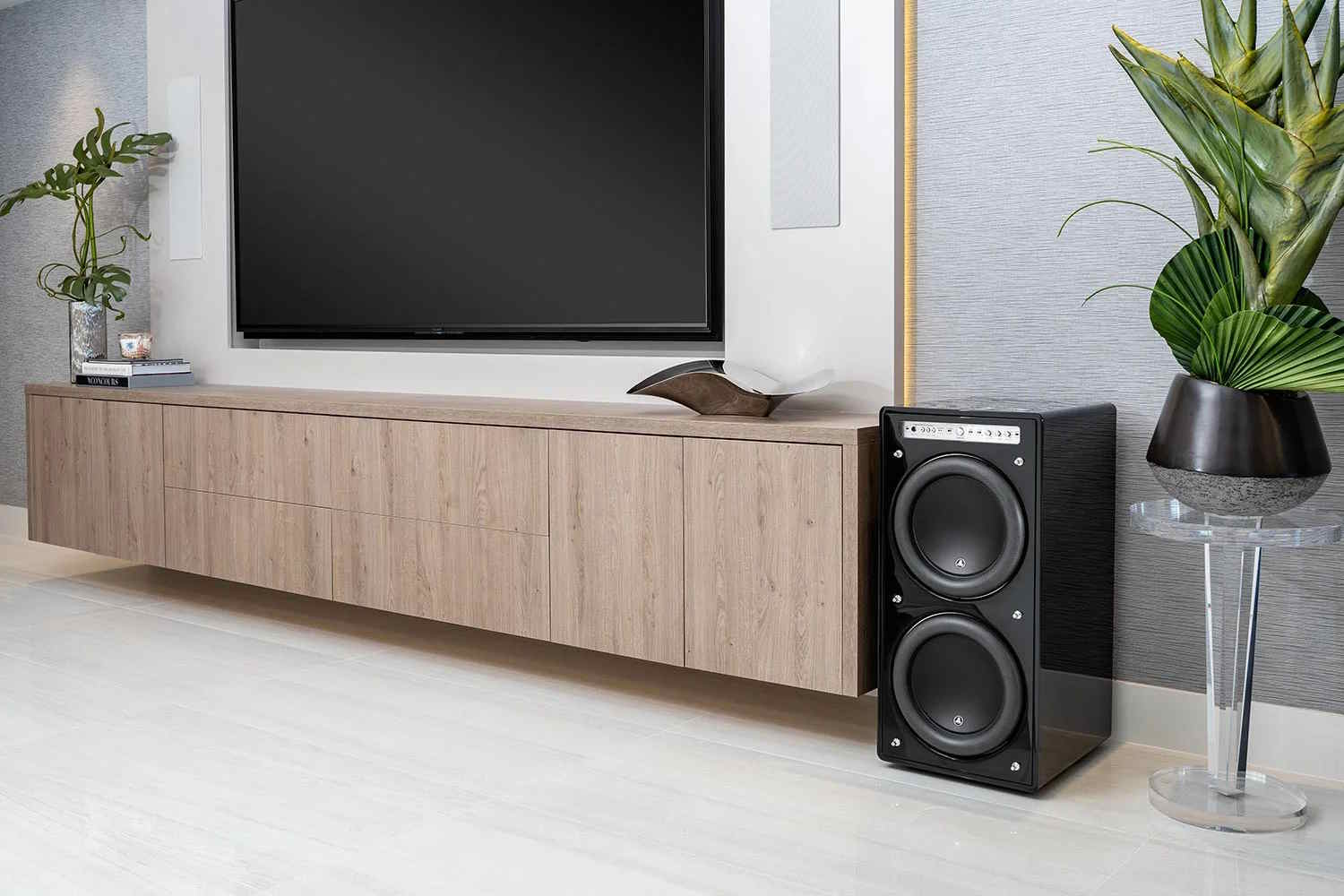 Modern living room entertainment setup featuring a wall-mounted TV above a floating wooden console, complemented by a large gloss-black subwoofer and green potted plants.