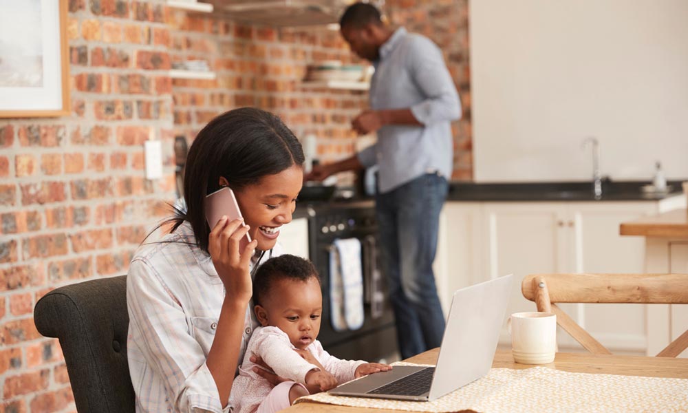 A mother smiles while talking on her smartphone and holding her baby at a dining table with a laptop, as a man cooks in the background kitchen.