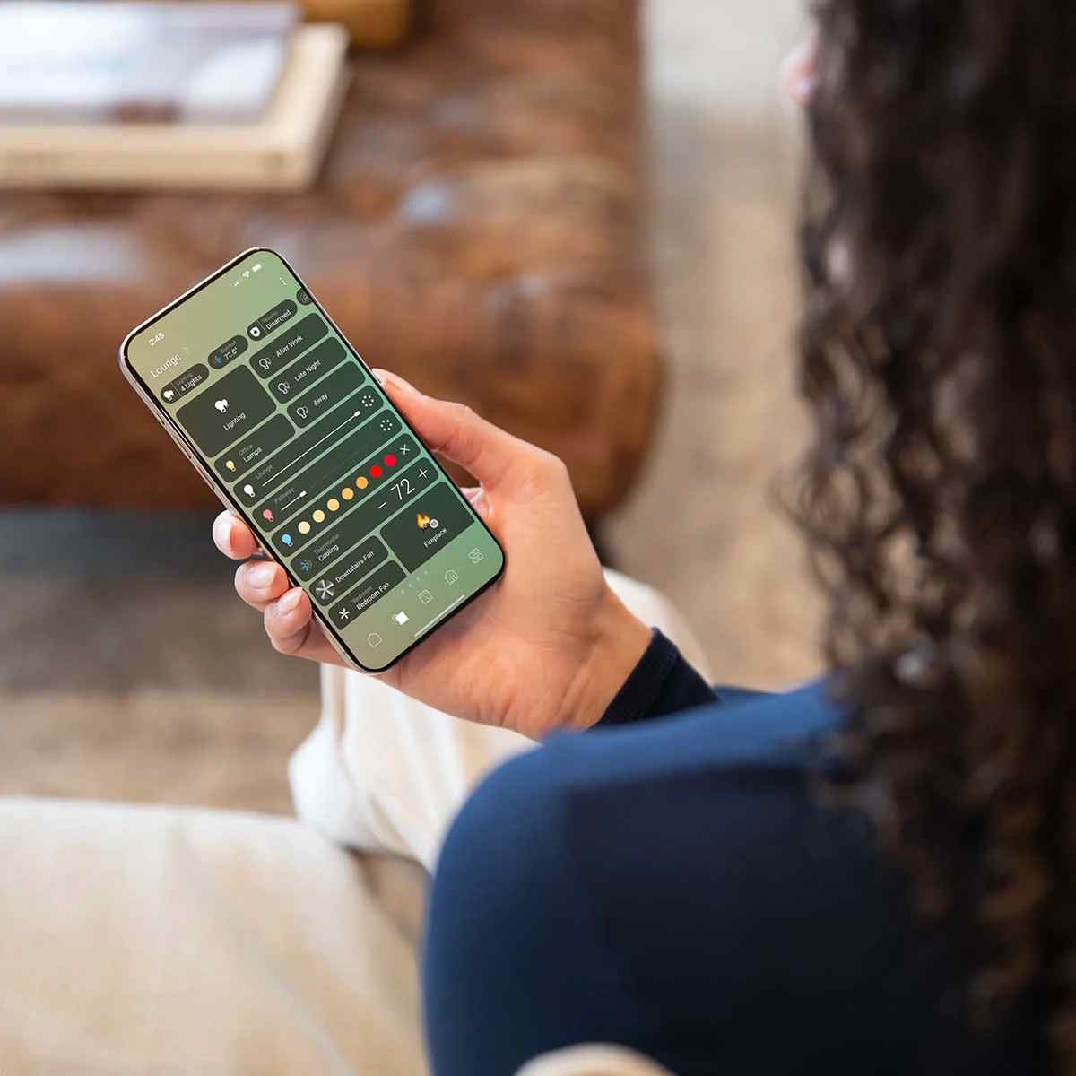 A person holds a smartphone displaying a smart home app interface with green tiles to control indoor lighting, fans, and home climate settings.