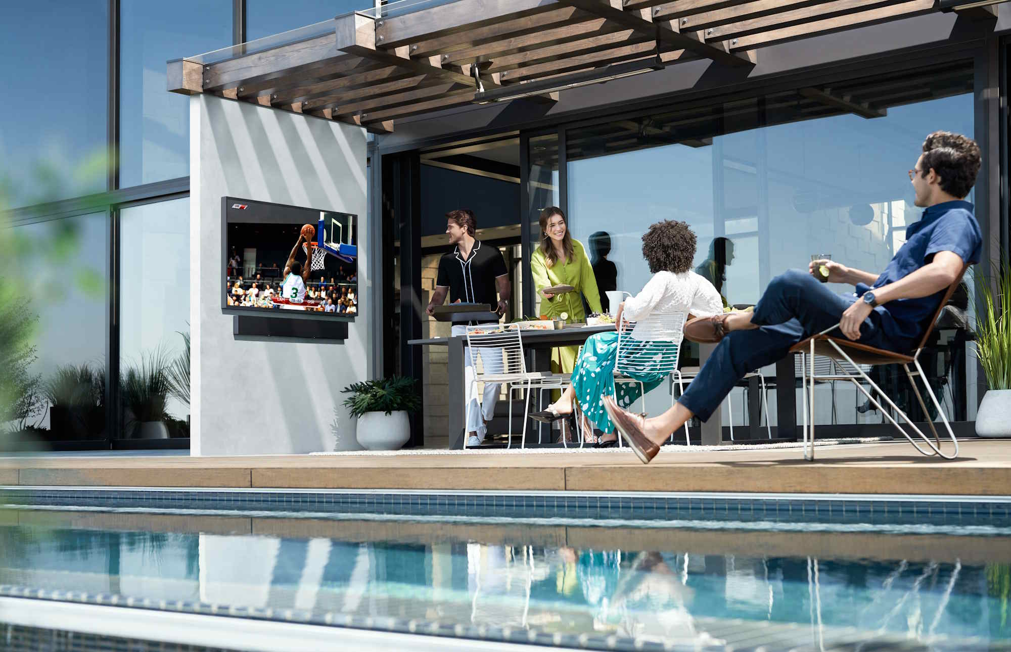 Outdoor patio with a pool, a pergola, and four people dining while a basketball game plays on a wall‑mounted TV.