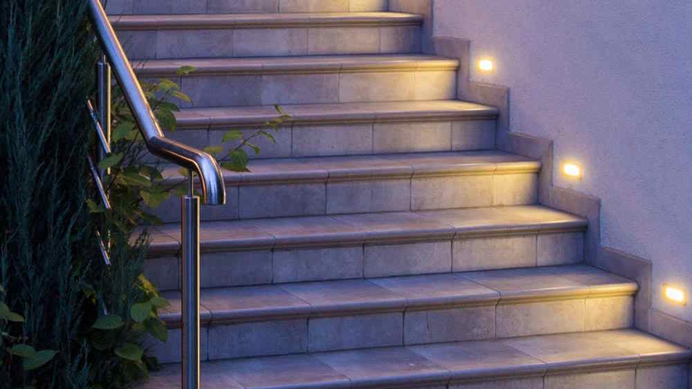 Outdoor stairs with warm recessed lighting and a metal handrail beside greenery.