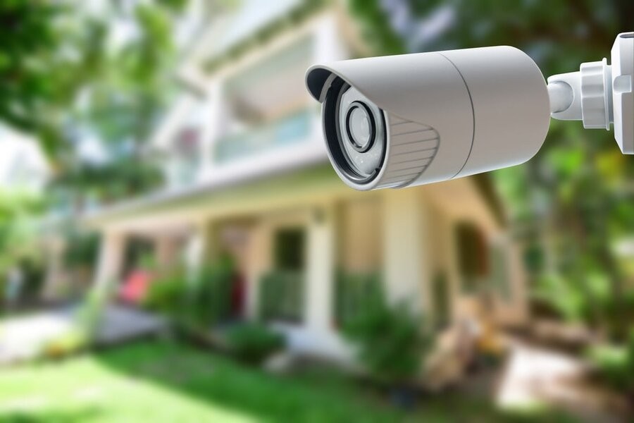 A smart home security camera in the foreground, with a house’s exterior in the background.