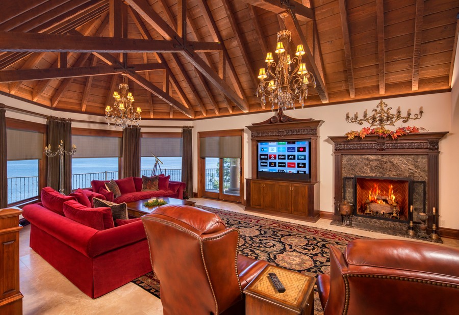 A living room with a wooden beam ceiling, motorized shades on windows, a fireplace, and TV in a cabinet. 