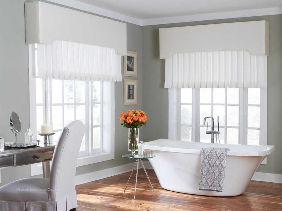 A bathroom with a soaking tub, vanity table, and chair. Lutron’s Roman shades are partially lowered over the windows.