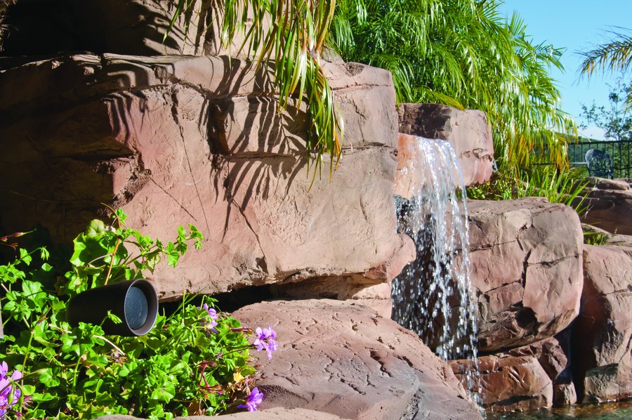 A small waterfall from boulders with a Sonance landscape speaker beside it amidst foliage.