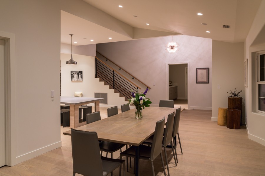Modern dining room with wood flooring, recessed ceiling lights, and a central wooden table surrounded by gray chairs, lit with warm ambient lighting at night.