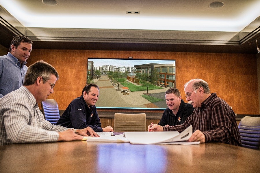 Contractors in a meeting room review plans with a concept drawing in the background.